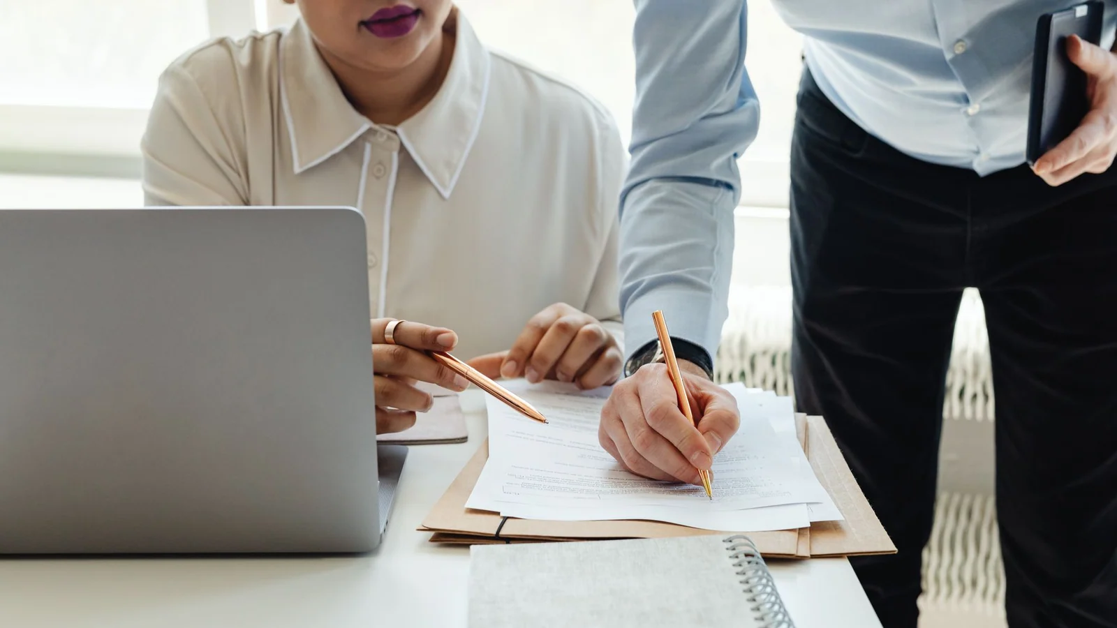 Two coworkers reviewing paperwork at a desk beside an open laptop