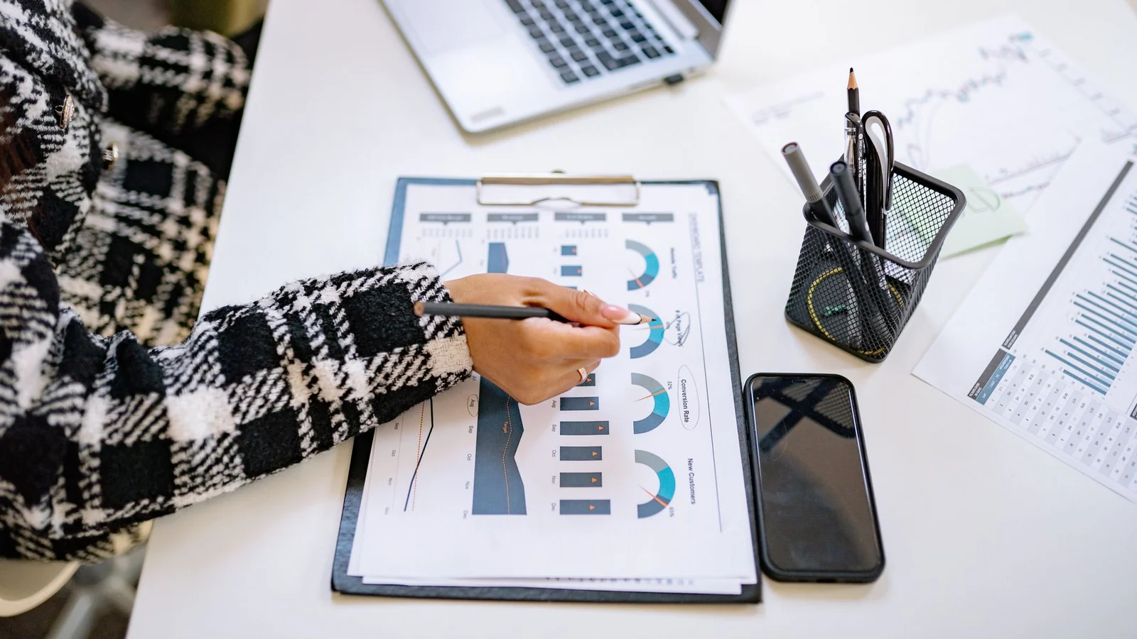 Hands marking a printed analytics report with charts beside a phone and laptop